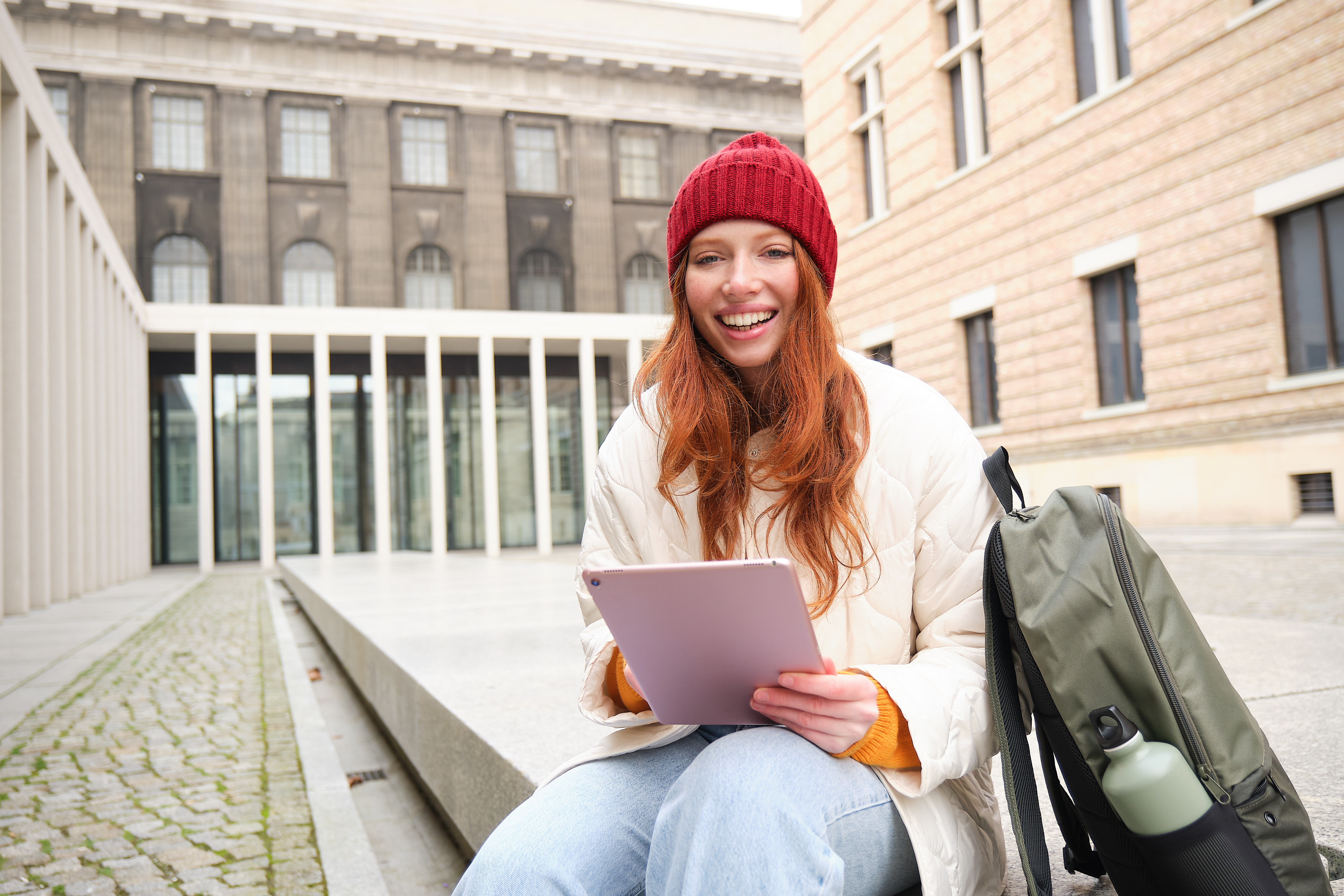 Beautiful redhead woman in red hat, sits with backpack and thermos, using digital tablet outdoors, connects to wifi, texts message, books tickets online.