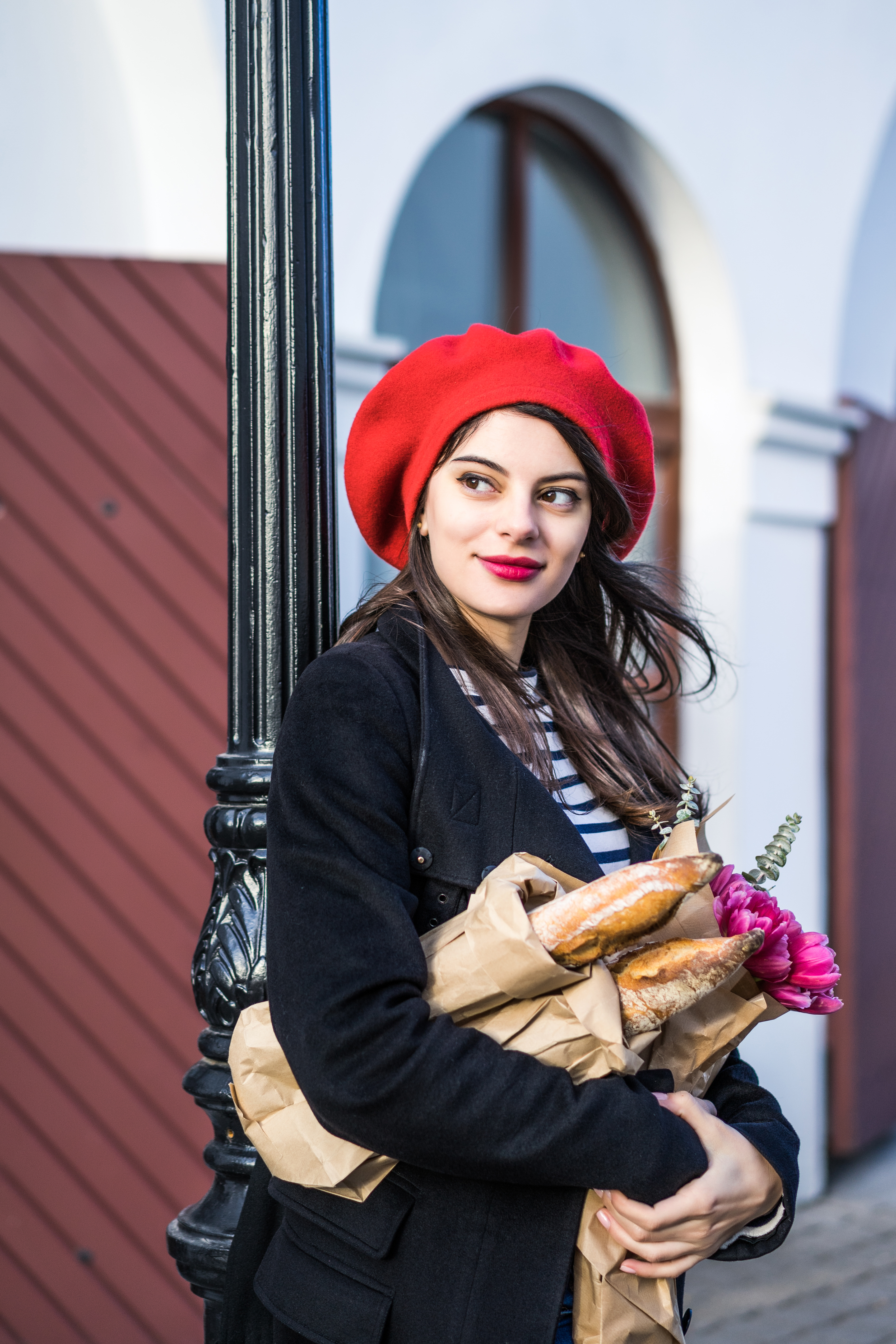 Young beautiful girl Frenchwoman brunette in a red beret and a black coat goes along the street of the European city with a bouquet of flowers and French baguettes. A happy smile and a charming look, the French style of Paris