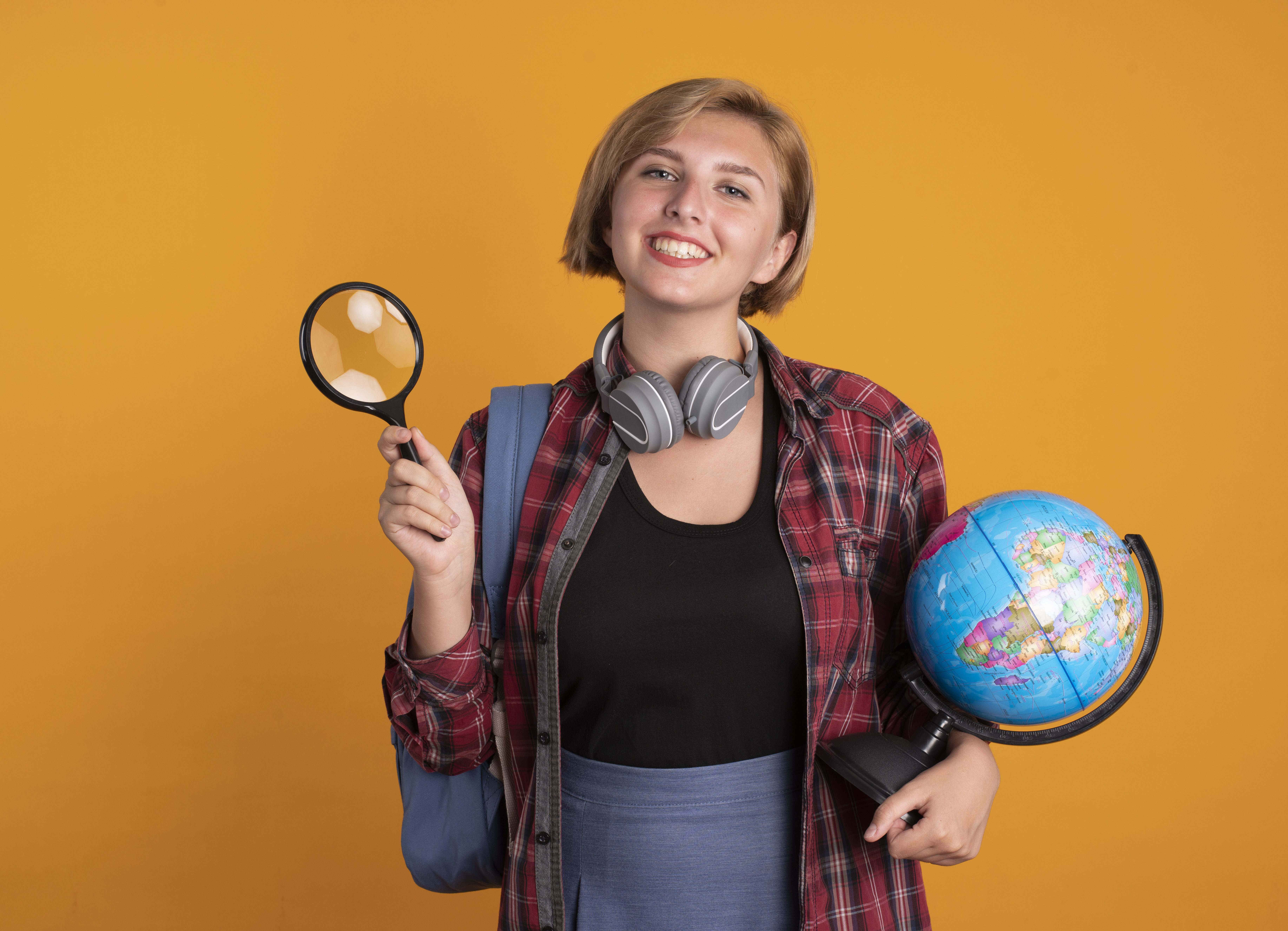 smiling young slavic student girl with headphones wearing backpack holds magnifying glass and globe isolated on orange background with copy space