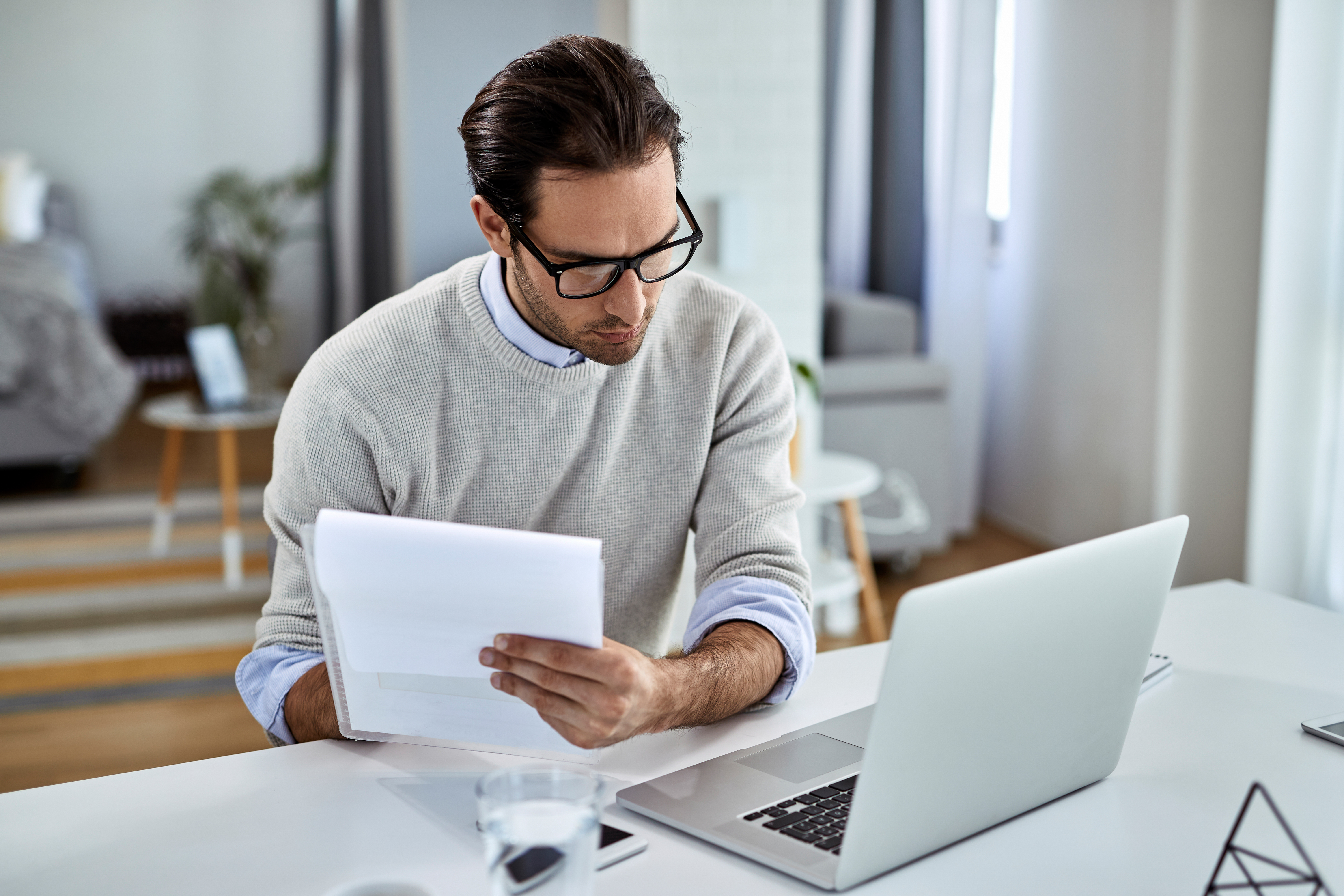 Young entrepreneur using laptop while working on business report Young businessman going through paperwork and using laptop while working at home.